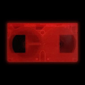 May include: A red VHS video cassette tape against a black background. The tape has a rectangular shape with two visible reel openings and a central hub. The plastic casing has a translucent quality, and the edges glow with a red light.