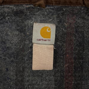 Puede incluir: Primer plano de una etiqueta Carhartt, con un logotipo de media luna amarilla sobre fondo blanco y el nombre de la marca en rojo oscuro. La etiqueta est&aacute; cosida sobre una tela gris texturizada, con un ribete de pana marr&oacute;n.