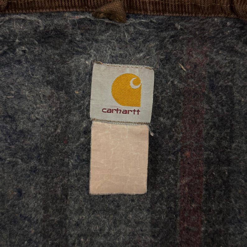 Puede incluir: Primer plano de una etiqueta Carhartt, con un logotipo de media luna amarilla sobre fondo blanco y el nombre de la marca en rojo oscuro. La etiqueta est&aacute; cosida sobre una tela gris texturizada, con un ribete de pana marr&oacute;n.