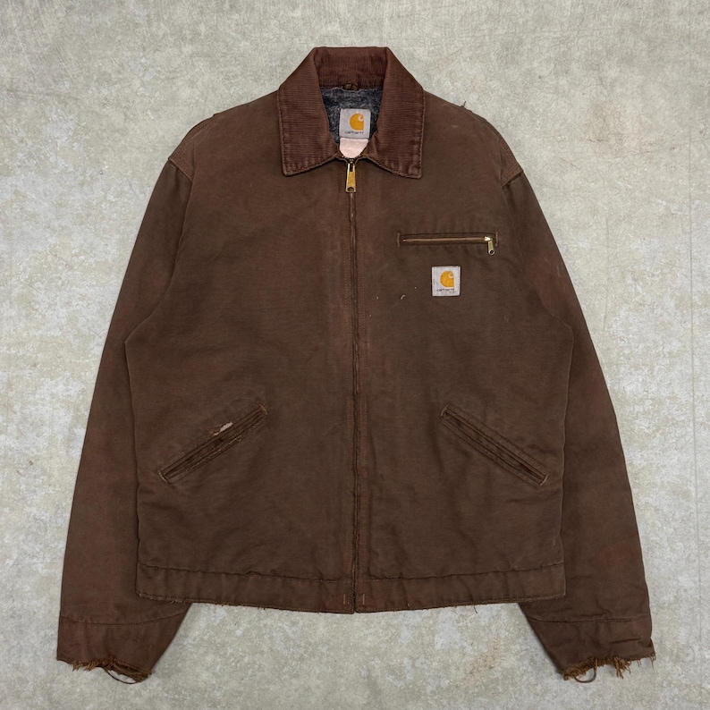 Puede incluir: Chaqueta de trabajo marr&oacute;n con cuello de pana, cierre de cremallera y m&uacute;ltiples bolsillos. La chaqueta presenta el logotipo de Carhartt en el bolsillo del pecho. La chaqueta est&aacute; hecha de un material duradero y desgastado.