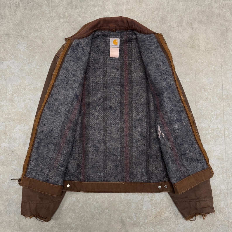 Puede incluir: Chaqueta de trabajo marr&oacute;n con cuello de pana y forro interior gris con estampado de cuadros. La chaqueta tiene cierre de botones y una peque&ntilde;a etiqueta con el logotipo. La chaqueta est&aacute; hecha de un material duradero y resistente.