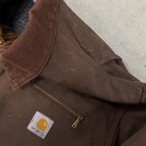 Puede incluir: Primer plano de una chaqueta Carhartt marr&oacute;n. La chaqueta presenta un cuello de pana, un bolsillo con cremallera y el parche con el logotipo de Carhartt. La chaqueta tiene un aspecto desgastado con algo de desgaste visible.
