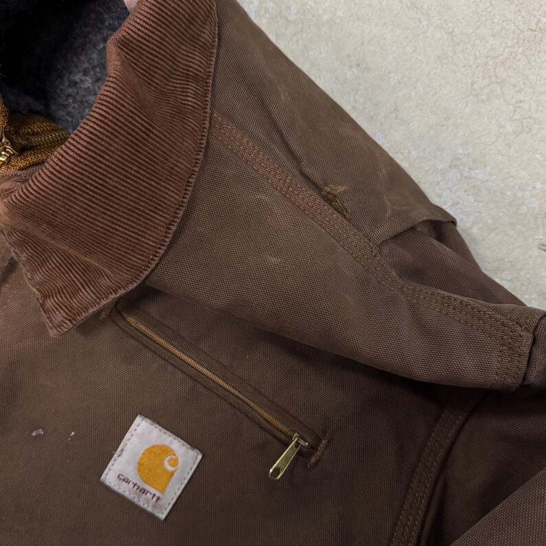 Puede incluir: Primer plano de una chaqueta Carhartt marr&oacute;n. La chaqueta presenta un cuello de pana, un bolsillo con cremallera y el parche con el logotipo de Carhartt. La chaqueta tiene un aspecto desgastado con algo de desgaste visible.