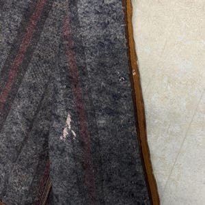 Puede incluir: Primer plano de una manta de lana gris con dos rayas burdeos. La manta tiene una cremallera marr&oacute;n y algo de desgaste. La manta es probablemente vintage o antigua.