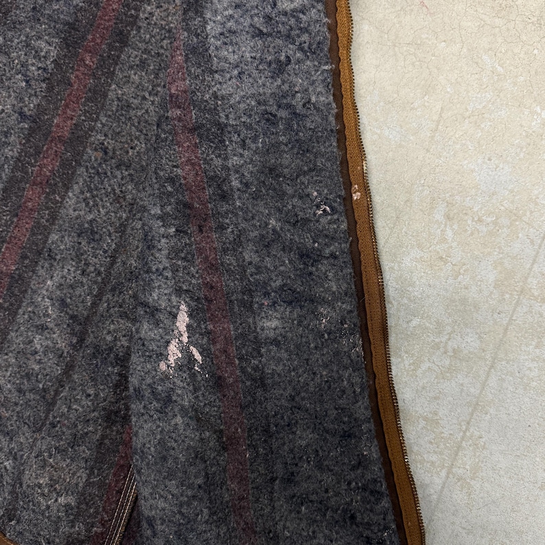 Puede incluir: Primer plano de una manta de lana gris con dos rayas burdeos. La manta tiene una cremallera marr&oacute;n y algo de desgaste. La manta es probablemente vintage o antigua.