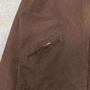 Puede incluir: Primer plano de una chaqueta marr&oacute;n con un bolsillo. La chaqueta muestra signos de desgaste, incluido un peque&ntilde;o agujero en el bolsillo. La tela parece ser un material duradero, posiblemente de trabajo. La est&eacute;tica general sugiere una prenda vintage o usada.