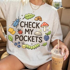 May include: A light grey t-shirt featuring the phrase "CHECK MY POCKETS" with cartoon illustrations of a blue beetle, ladybug, mushroom, caterpillar, and other nature-themed graphics.