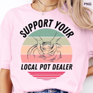 May include: A light pink t-shirt featuring a graphic of hands shaping clay on a pottery wheel. The design includes a multi-colored striped circle in green, peach, and red, with the black text SUPPORT YOUR LOCAL POT DEALER printed around it.