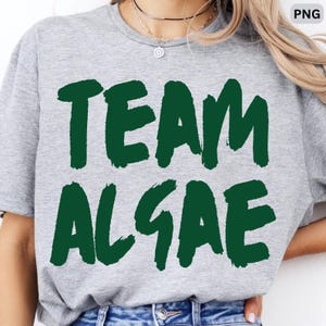 May include: A heather gray short-sleeve t-shirt featuring the text TEAM ALGAE printed in a bold, dark green, brush-stroke style font across the chest. The shirt has a classic crew neckline and a standard fit, providing a casual look.