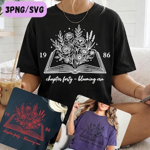Può includere: T-shirt nero con un design floreale bianco con rose, margherite e lavanda, sopra un libro aperto. Il testo include "19 86" e "chapter forty - blooming era". Sono mostrate anche magliette aggiuntive in blu navy e viola.