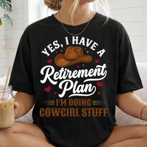 May include: Black t-shirt with the text "YES, I HAVE A Retirement Plan" above a brown cowboy hat graphic. Below the hat, the text reads "I'M DOING COWGIRL STUFF". Red heart graphics are scattered around the text.