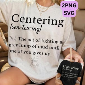 Op de afbeelding: Lichtbeige t-shirt met het woord "Centering" in grote zwarte letters, gevolgd door een fonetische uitspraak en een definitie. De definitie luidt: "De handeling van het bestrijden van een grijze klomp modder totdat een van jullie opgeeft."
