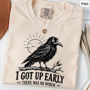 Könnte beinhalten: Ein cremefarbenes T-Shirt mit einer schwarzen Grafik eines Raben, der auf einem Ast vor einer Sonne sitzt. Unter dem Raben steht der Text I GOT UP EARLY THERE WAS NO WORM. Das T-Shirt hat ein Comfort Colors Markenetikett am inneren Halsausschnitt.