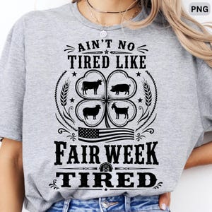 Può includere: Una t-shirt grigio melange con testo nero che recita AIN'T NO TIRED LIKE FAIR WEEK TIRED. Il design include un quadrifoglio contenente le sagome di una mucca, un maiale, una pecora e una capra, insieme a una piccola grafica della bandiera americana.
