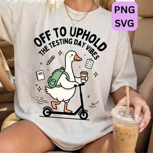 Può includere: T-shirt beige con grafica di un'oca su uno scooter, con zaino e caffè, con la scritta "OFF TO UPHOLD THE TESTING DAY VIBES". Il design include stelle, una matita, un taccuino e un documento.