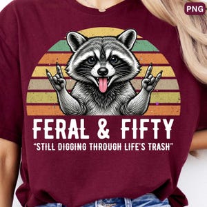 Puede incluir: Una camiseta color granate con el gráfico de un mapache con la lengua fuera haciendo un gesto de rock-and-roll. Detrás hay un atardecer retro en amarillo, naranja y verde. El texto dice FERAL & FIFTY STILL DIGGING THROUGH LIFE'S TRASH.