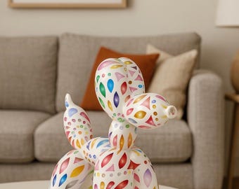 May include: A white ceramic balloon dog sculpture with colorful abstract patterns in red, blue, yellow, and green. The sculpture is on a white table, with a sofa and artwork in the background. Home decor.