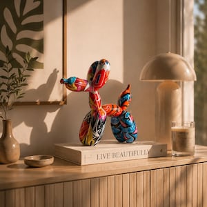 Peut inclure: Une sculpture de chien ballon colorée avec un motif abstrait vibrant en rouge, bleu, jaune et noir. La sculpture est posée sur un livre avec l'inscription "LIVE BEAUTIFULLY". Un petit bol, un vase et une bougie sont également sur la surface en bois.