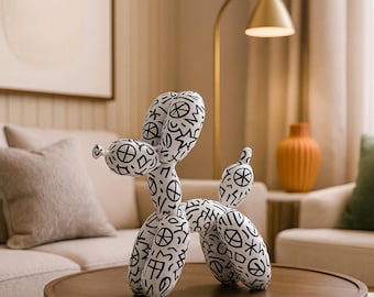 May include: A fabric balloon dog sculpture with a repeating black and white pattern, placed on a dark wooden coffee table. The background features a beige sofa, artwork, and a gold-coloured lamp. The sculpture is a decorative item.