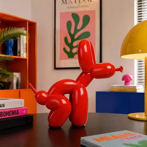 Peut inclure: Une sculpture rouge brillante en forme de chien ballon en position debout. La figurine présente des segments arrondis évoquant des ballons gonflés formant la tête, les oreilles, le corps et les pattes, avec une petite queue et un museau noué.