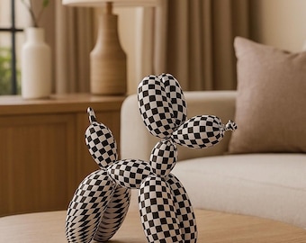 May include: A black and white checkered balloon dog sculpture. The dog is posed on a round wooden table. The sculpture is a decorative item, resembling a balloon animal. The background includes a lamp, sofa, and curtains.