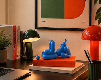 Op de afbeelding: Een blauwe ballonsculptuur van een liggende figuur op een oranje boek getiteld Dieter Rams: The Complete Works. Het beeldje staat tussen een donkergroene paddenstoelvormige lamp en een feloranje paddenstoelvormige lamp in.