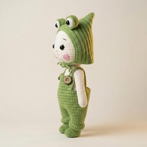以下が含まれることがあります： 手作りの編み人形、緑色のカエルのコスチュームを着ています。人形は白い顔にピンクの頬、大きな黒い目があります。カエルの目がある緑色の帽子と、木製のボタンが付いた緑色のオーバーオールを着ています。
