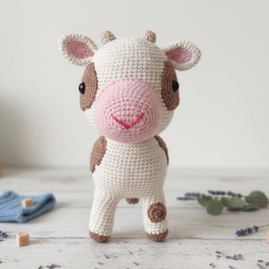 Puede incluir: Un juguete de vaca hecho a mano con ganchillo. La vaca es principalmente de color blanco roto con manchas y detalles marrones. Tiene una nariz y boca rosas, ojos negros y pequeños cuernos. El juguete está de pie, mostrando el detallado trabajo de ganchillo.