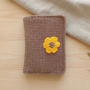 Könnte beinhalten: Eine braune gehäkelte Hülle mit einer Applikation aus gelben und goldenen Blumen. Die Hülle ist rechteckig und scheint handgefertigt zu sein. Die Blume ist auf der Vorderseite der Hülle zentriert.