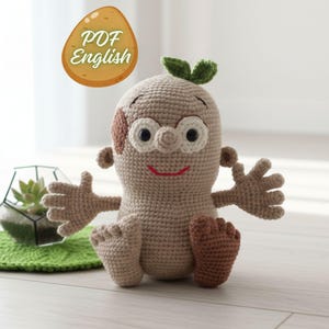 Puede incluir: Muñeco de ganchillo hecho a mano en color marrón claro, con una cara sonriente, ojos negros, boca roja y una pequeña mancha marrón. Tiene brazos y pies pequeños, y una hoja verde en la cabeza. La imagen incluye el texto "PDF English".