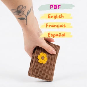 May include: A brown crocheted wallet with a yellow flower applique. The wallet is held in a hand, with text that reads "PDF", "English", "Français", and "Español" in the background.