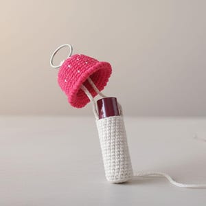 May include: A handmade crochet lipstick holder featuring a pink mushroom cap with white spots. The holder is white, with a matching white cord. A silver ring is attached to the mushroom cap. The lipstick is visible.