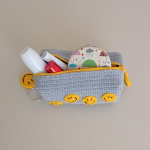May include: A gray crochet cosmetic bag with a yellow zipper and smiley face accents. The bag contains white and red cosmetic items and a small patterned compact mirror. A yellow smiley face keychain is attached.