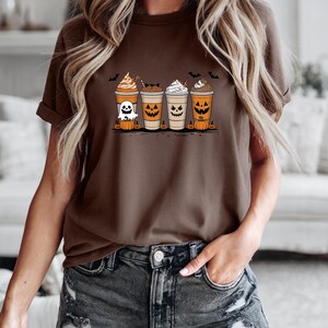 May include: A brown t-shirt featuring a Halloween-themed graphic. The graphic depicts four coffee cups with spooky faces, whipped cream, and pumpkins. Bats are flying above the cups. The shirt is worn by a person.
