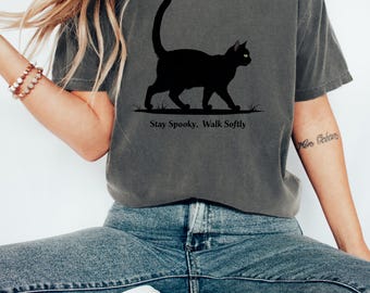 May include: Dark grey t-shirt featuring a black cat silhouette walking on grass. The text "Stay Spooky. Walk Softly" is printed below the cat. The shirt is made of a soft material and has a relaxed fit.