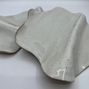 May include: Two off-white ceramic plates with a speckled surface and wavy edges. The plates have a rustic, handmade appearance, with a slightly rough texture and a warm brown rim. The plates are designed for serving food.