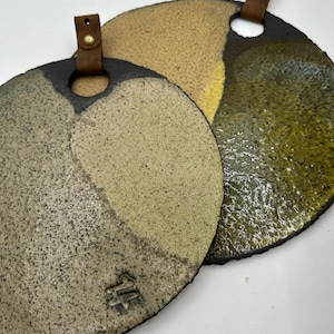 May include: Two round, ceramic serving platters with a speckled, textured surface in shades of tan, olive green, and black. Each platter has a leather strap attached with a brass rivet for hanging. The platters are handmade.