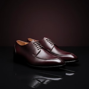 May include: A pair of burgundy leather dress shoes with a textured finish. The shoes have a classic lace-up design and a sleek, polished appearance. The shoes are displayed on a reflective black surface.