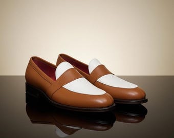 May include: A pair of brown leather loafers with white accents on the vamp and a red interior. The shoes are displayed on a reflective black surface, with a neutral background.