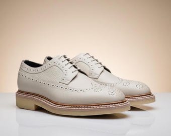 May include: A pair of off-white leather brogue shoes with a textured finish. The shoes feature a classic wingtip design with decorative perforations and a light brown sole. The laces are white, and the shoes are set against a neutral background.