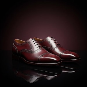 Könnte beinhalten: Ein Paar burgunderfarbene Lederschuhe mit schwarzen Schnürsenkeln. Die Schuhe haben ein Cap-Toe-Design und dekorative Perforationen entlang der Nähte. Die Schuhe werden auf einer reflektierenden Oberfläche präsentiert.