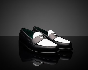 May include: A pair of black and white leather penny loafers. The shoes have a classic design with a black upper, white vamp, and a green interior lining. The shoes are displayed on a reflective surface.