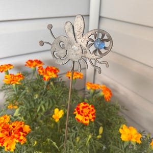 May include: A metal bee garden stake with a blue gem eye, set amidst vibrant orange and yellow marigold flowers. The bee has a whimsical design and is placed in a pot of green foliage.