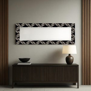 Può includere: Uno specchio rettangolare con una cornice nera con un motivo a foglie bianche ripetute. Lo specchio è sopra un mobile in legno marrone scuro con scanalature verticali. Una lampada con un paralume beige è sul mobile.