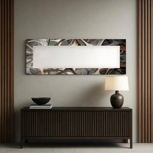 May include: A rectangular wall mirror with a white center and an abstract, metallic-toned frame. The mirror hangs above a dark brown wooden cabinet with vertical grooves. A black bowl and a lamp with a brown base and cream shade are also visible.