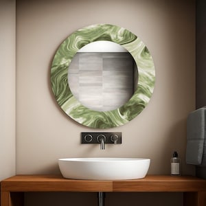 May include: A round mirror with a green marble-like frame hangs above a white oval sink. The mirror reflects a tiled wall. The sink sits on a wooden vanity with a black faucet and a soap dispenser.