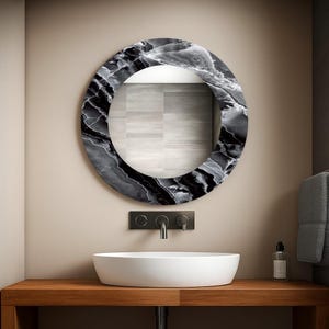 Peut inclure: Un miroir de salle de bain rond avec un cadre noir et blanc effet marbre. Le miroir est au-dessus d'un lavabo blanc ovale sur un comptoir en bois. Un robinet noir et des accessoires muraux sont visibles.