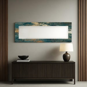 May include: A rectangular wall mirror with a teal, gold, and white textured frame hangs above a dark brown cabinet. A black bowl and a lamp with a brown base and white shade sit on the cabinet.