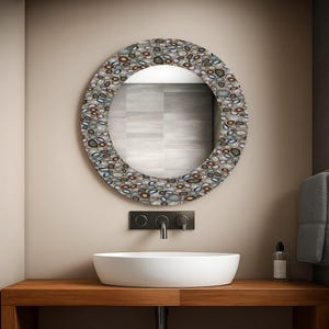 May include: A round mirror with a decorative border featuring a pebble design in shades of gray, brown, and blue. The mirror is mounted above a white, oval-shaped sink and a wooden countertop in a bathroom setting.
