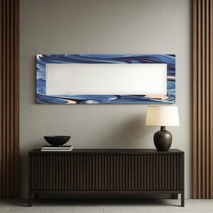 May include: A rectangular mirror with a blue and white abstract design frame hangs above a dark brown wooden cabinet. A black bowl and books sit on the cabinet. A brown lamp with a cream shade is on the cabinet.